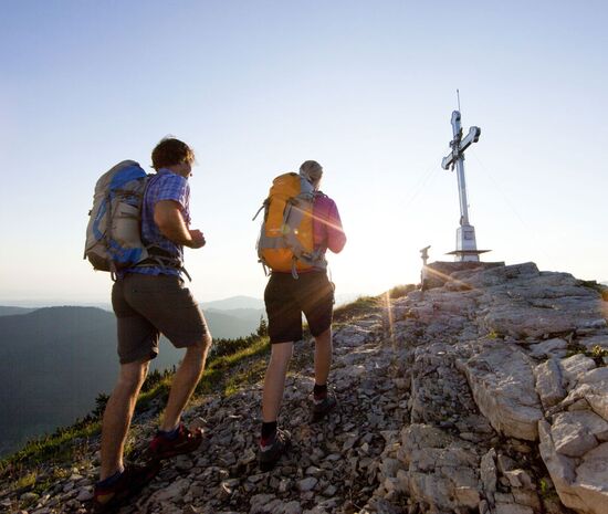 2 hikers stand at the summit cross of the Wallberg at sunrise