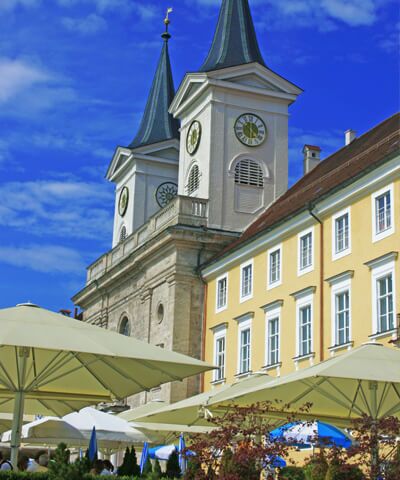 Blick auf die Schlosskirche Tegernsee bei blauem Himmel vor der Schlosskirche ist der Freisitz mit Tischen Stuehlen und geoeffneten Sonnenschirmen des Brauhaus Tegernsee